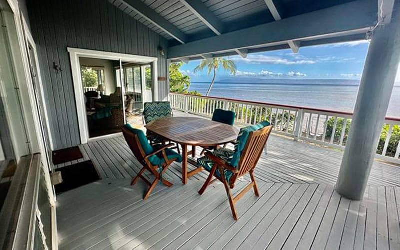 A covered lanai on a Molokaʻi home, with a wooden table and chairs on a painted deck, looking past a palm tree and a white railing to the ocean and a distant island on the horizon