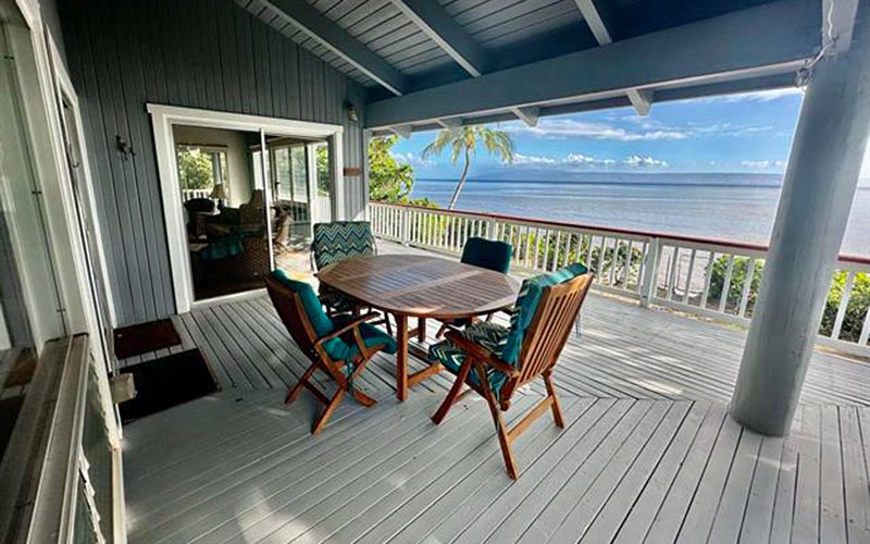 A covered lanai on a Molokaʻi home, with a wooden table and chairs on a painted deck, looking past a palm tree and a white railing to the ocean and a distant island on the horizon