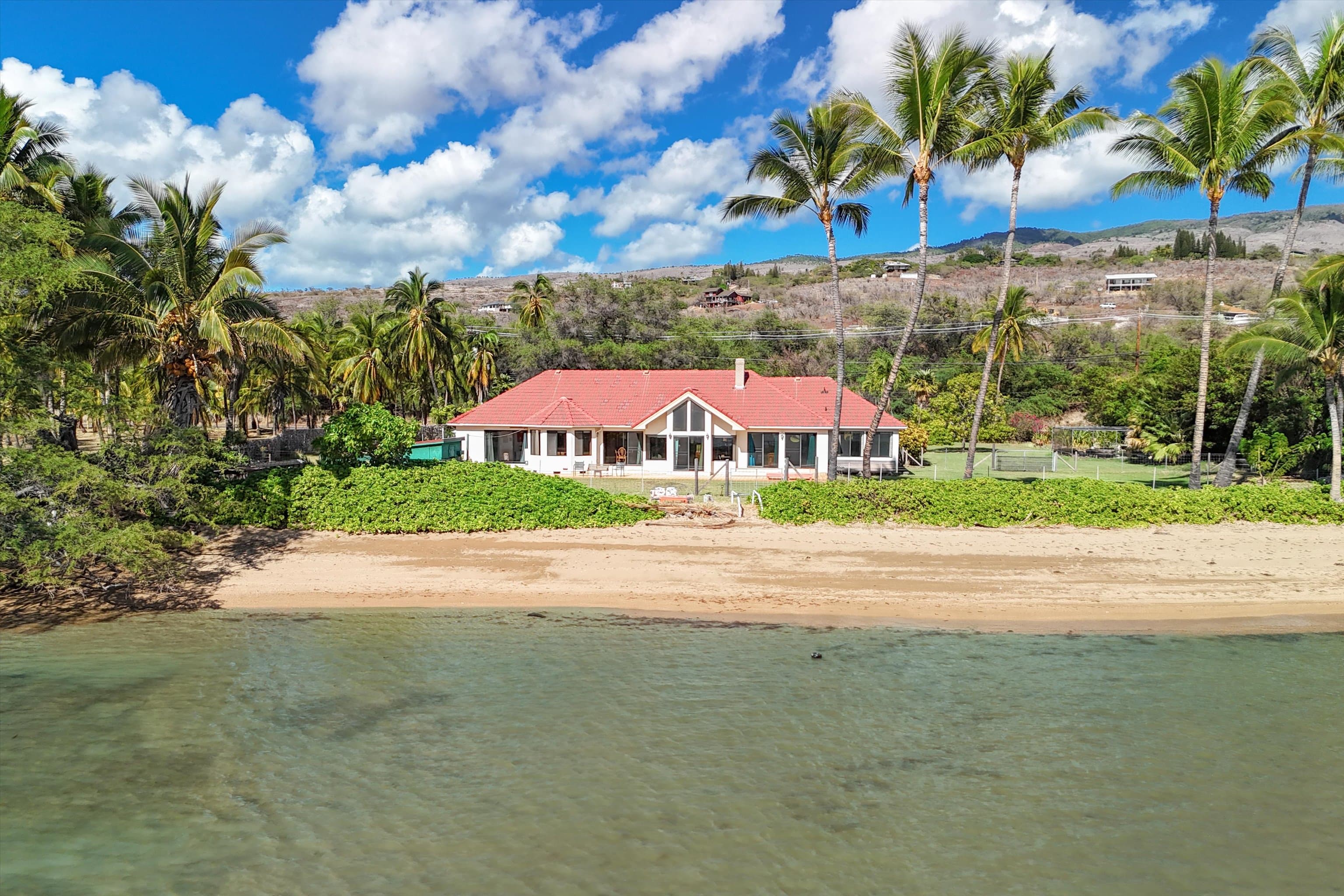 A red-roofed oceanfront home on a Molokaʻi beach, framed by tall palms, with the dry hillside and scattered houses rising behind it