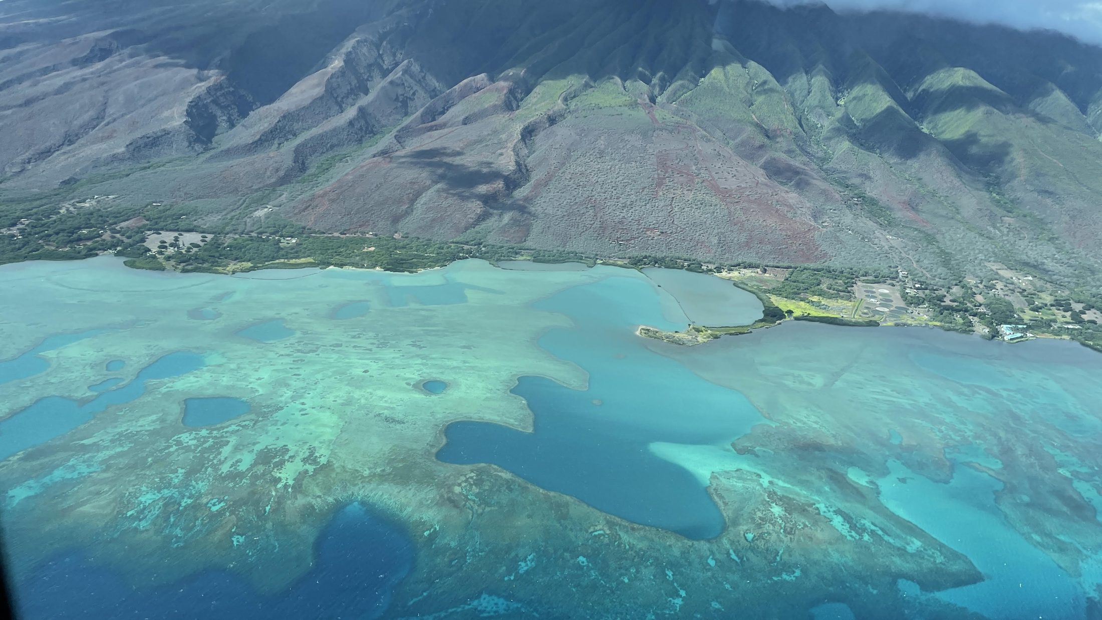 Aerial view of Molokaʻi's lush green mountains rising above a vast turquoise coral reef and calm Pacific shallows