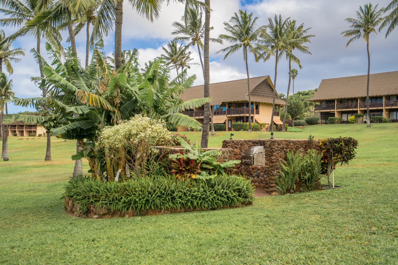 A stone barbecue pavilion and tropical planting on the resort's open lawn, with cedar-shingled two-storey condominium buildings and palms behind