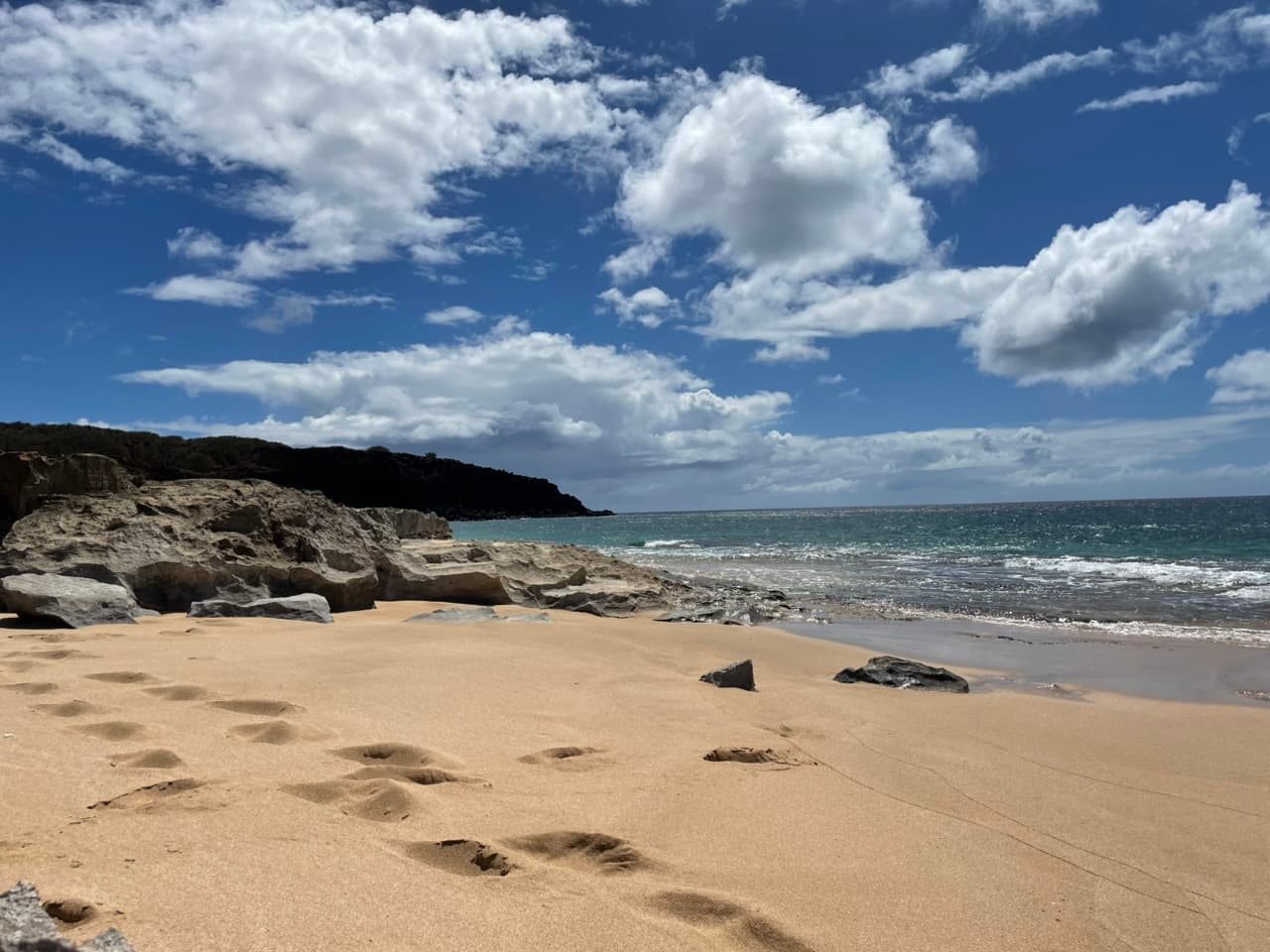 Kepuhi Beach looking along the shore, with weathered rock formations, footprints in the sand and open ocean
